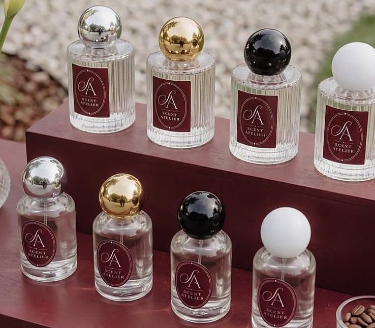 Osam bočica Scent Atelier na oxblood baru — dva oblika stakla, svaki sa srebrnim, zlatnim, crnim i bijelim čepom.